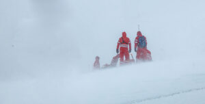 Mountain rescue team in red cold-weather gear assisting an injured person during a whiteout snowstorm, illustrating the question Do I need insurance for my hunting trip in remote wilderness conditions.