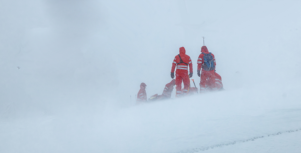 Mountain rescue team in red cold-weather gear assisting an injured person during a whiteout snowstorm, illustrating the question Do I need insurance for my hunting trip in remote wilderness conditions.